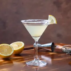 Klasyczny martini z cytryną i solą na kieliszku. W tle cytryny, oliwki i akcesoria barmańskie. Poznaj różne martini rodzaje.