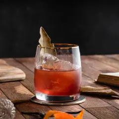 Elegancki koktajl, prawdopodobnie martini extra dry, z lodem i liściem laurowym, na drewnianym stole obok starej książki.