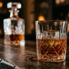 Szklanka z lodem i whisky na drewnianym blacie. W tle karafka, sugerująca różne rodzaje whisky.
