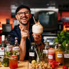 Uśmiechnięty barman z koktajlem w dłoni, otoczony butelkami alkoholi, w tym Bacardi. Sprawdź aktualną bacardi cena!