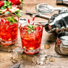 Dwa orzeźwiające drinki z martini rosato, lodem i truskawkami, obok shakera i akcesoriów barmańskich.