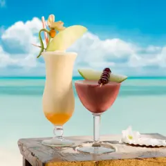 Dwa tropikalne drinki, jeden kremowy, drugi czerwony, ozdobione owocami i kwiatami, stoją na drewnianym stoliku na plaży. Idealne na upalny dzień, jak ten z **malibu alkohol**.