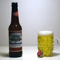 Butelka i kufel pełne złotego piwa Budweiser. Obok leży kapsel z napisem "Bud".