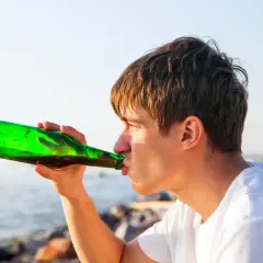 Młody mężczyzna pije piwo z zielonej butelki nad wodą. Słońce odbija się w jego włosach, tworząc ciepły blask.