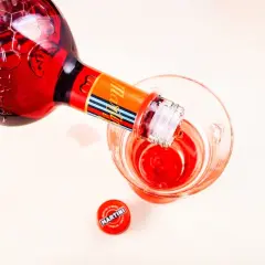 Czerwony Martini Fiero nalewany do kieliszka. Obok leży czerwona zakrętka z logo Martini.