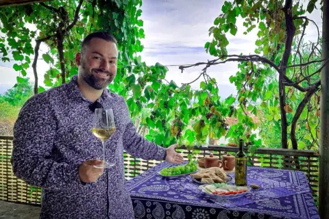 Mężczyzna z kieliszkiem białego wina, pod winoroślą, zaprasza do degustacji marani wino. Na stole owoce, chleb i ser.