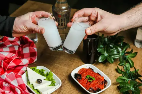 Dwie szklanki z mleczną wódką turecką (rakı) unoszą się do toastu. Na stole ser biały, oliwka i sos pomidorowy z bakłażanem.