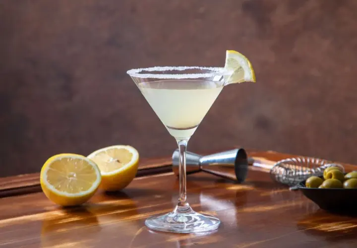 Klasyczny martini z cytryną i solą na kieliszku. W tle cytryny, oliwki i akcesoria barmańskie. Poznaj różne martini rodzaje.