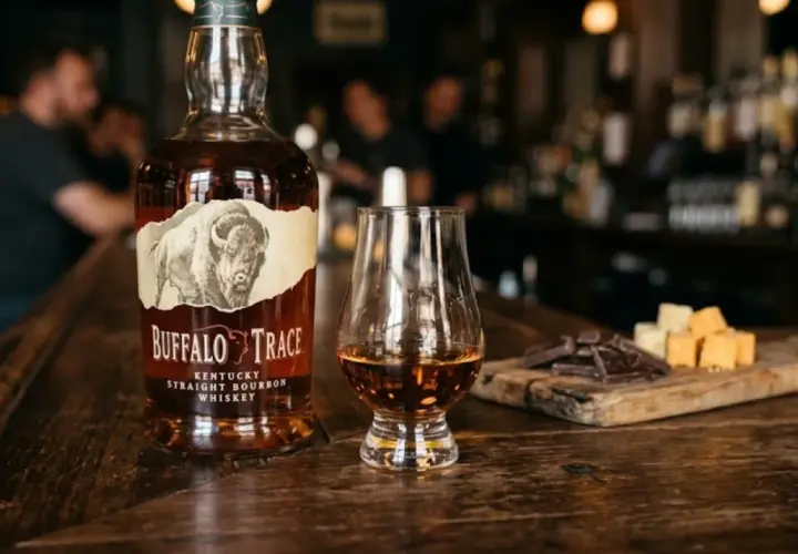 Butelka burbona Buffalo Trace, kieliszek z bursztynowym płynem, kawałki czekolady i sera na drewnianej desce.