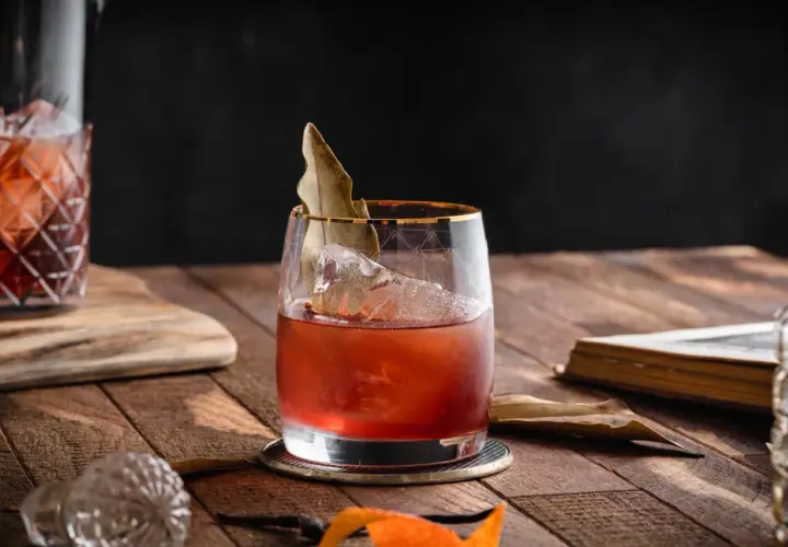 Elegancki koktajl, prawdopodobnie martini extra dry, z lodem i liściem laurowym, na drewnianym stole obok starej książki.