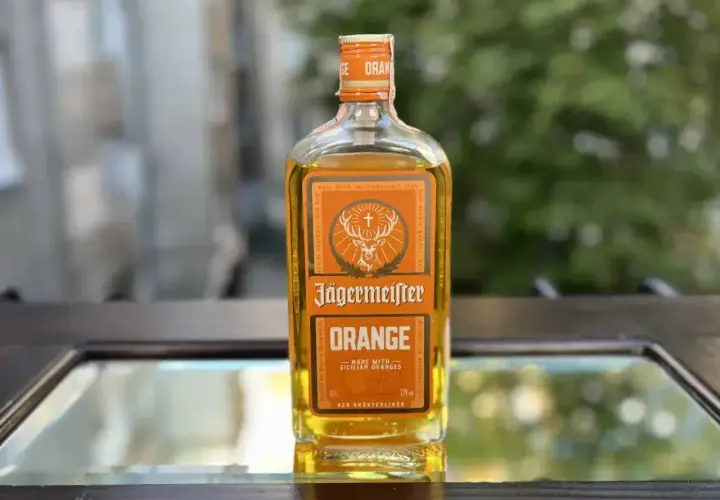 Butelka Jägermeister Orange, z pomarańczowym akcentem, stoi na szklanym blacie.