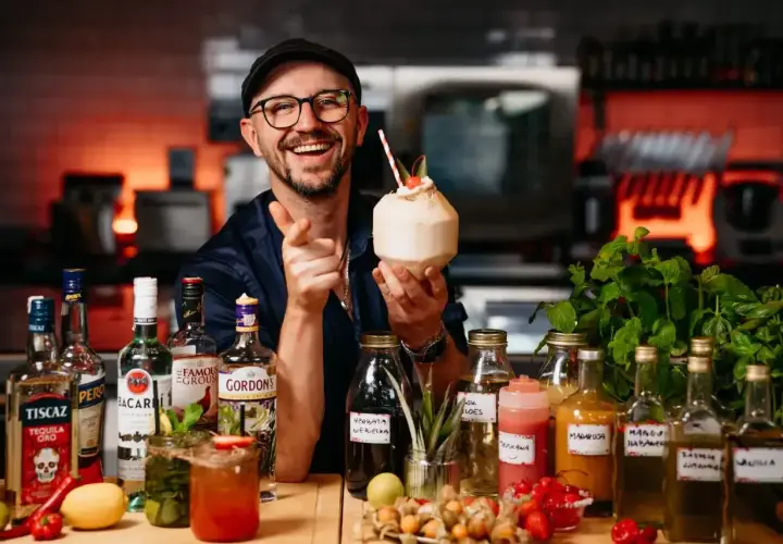 Uśmiechnięty barman z koktajlem w dłoni, otoczony butelkami alkoholi, w tym Bacardi. Sprawdź aktualną bacardi cena!