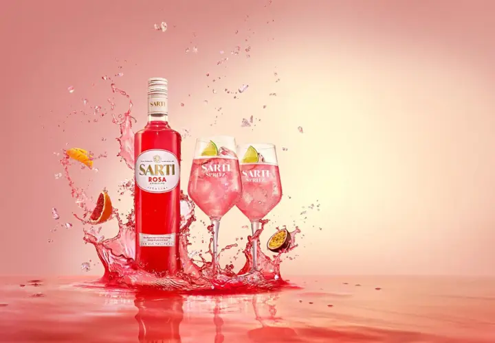 Sarti Rosa Aperitivo i dwa drinki Sarti Spritz z lodem i limonką, otoczone eksplozją owoców i różowego płynu.