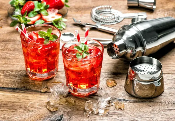 Dwa orzeźwiające drinki z martini rosato, lodem i truskawkami, obok shakera i akcesoriów barmańskich.