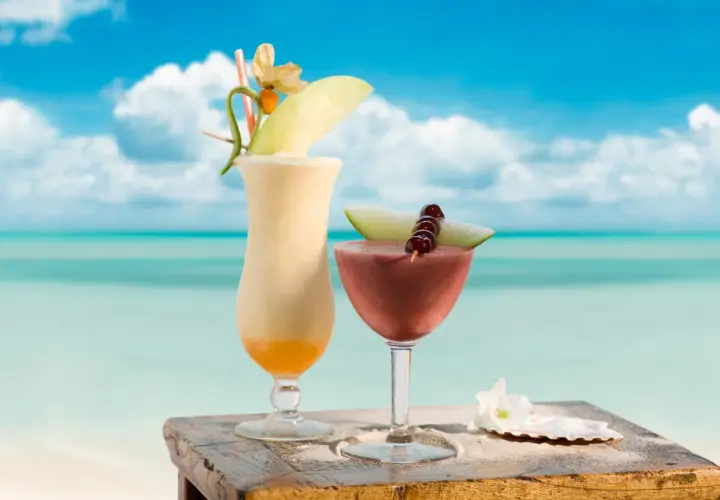Dwa tropikalne drinki, jeden kremowy, drugi czerwony, ozdobione owocami i kwiatami, stoją na drewnianym stoliku na plaży. Idealne na upalny dzień, jak ten z **malibu alkohol**.