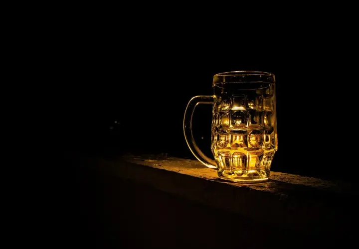 Złocisty kufel piwa Grimbergen, podświetlony od tyłu, stoi na kamiennym murku w ciemności.