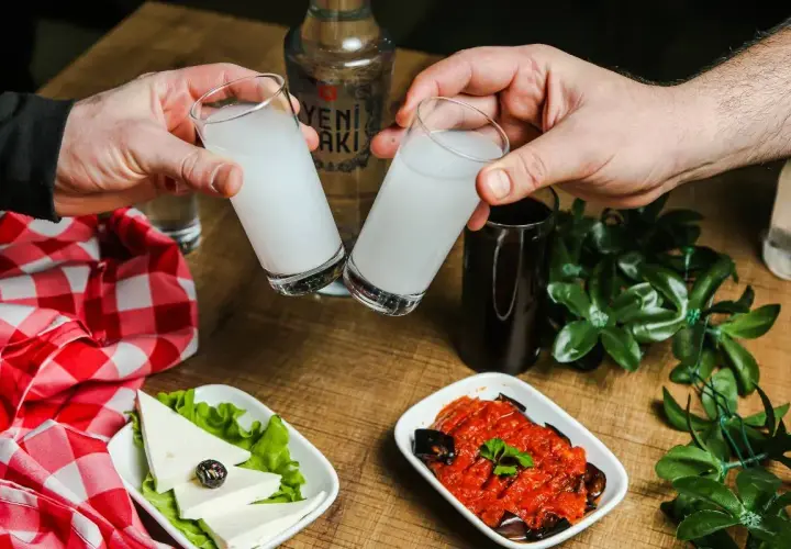 Dwie szklanki z mleczną wódką turecką (rakı) unoszą się do toastu. Na stole ser biały, oliwka i sos pomidorowy z bakłażanem.