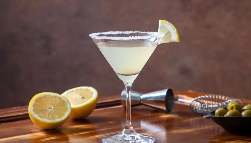 Klasyczny martini z cytryną i solą na kieliszku. W tle cytryny, oliwki i akcesoria barmańskie. Poznaj różne martini rodzaje.