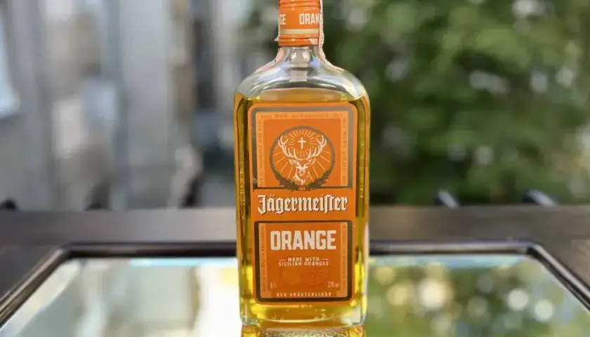 Butelka Jägermeister Orange, z pomarańczowym akcentem, stoi na szklanym blacie.