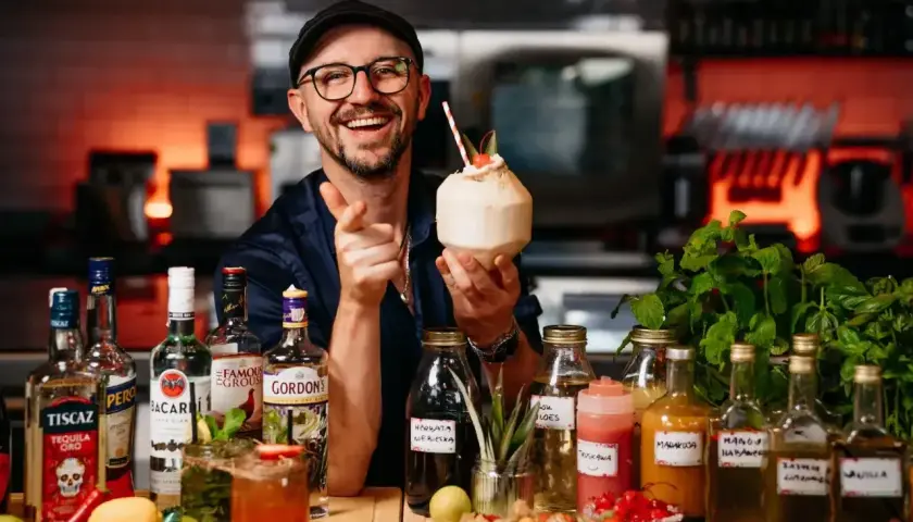 Uśmiechnięty barman z koktajlem w dłoni, otoczony butelkami alkoholi, w tym Bacardi. Sprawdź aktualną bacardi cena!