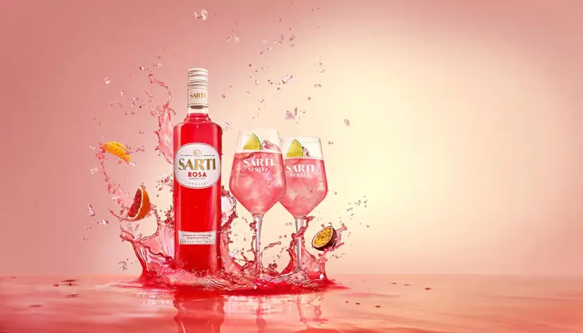 Sarti Rosa Aperitivo i dwa drinki Sarti Spritz z lodem i limonką, otoczone eksplozją owoców i różowego płynu.