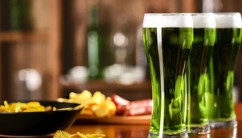 Trzy szklanki zielonego piwa z pianką, obok miska chipsów i kiełbasa. Idealne na świętowanie Dnia Świętego Patryka.