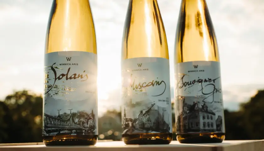 Trzy butelki białego wina Winnica Aris: Solaris, Muscat i Sauvignon Blanc, stojące na tle zachodzącego słońca.