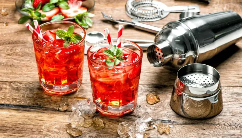 Dwa orzeźwiające drinki z martini rosato, lodem i truskawkami, obok shakera i akcesoriów barmańskich.