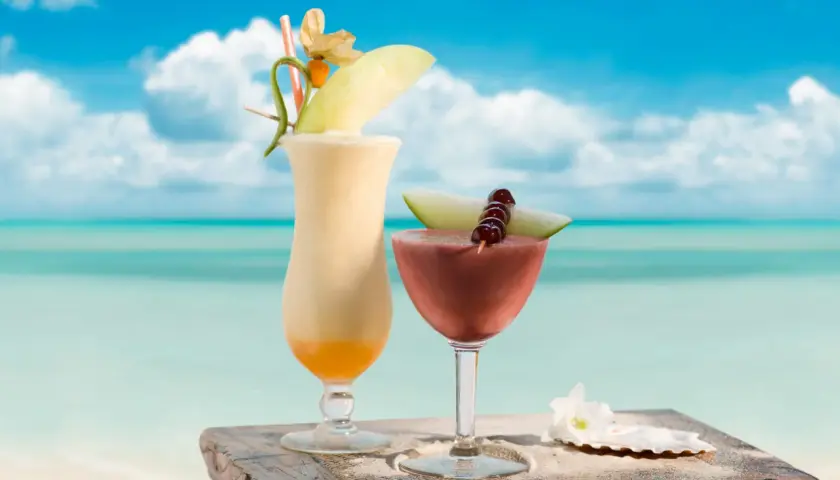 Dwa tropikalne drinki, jeden kremowy, drugi czerwony, ozdobione owocami i kwiatami, stoją na drewnianym stoliku na plaży. Idealne na upalny dzień, jak ten z **malibu alkohol**.