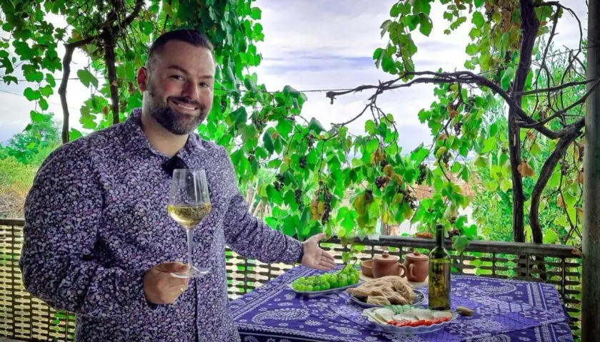Mężczyzna z kieliszkiem białego wina, pod winoroślą, zaprasza do degustacji marani wino. Na stole owoce, chleb i ser.