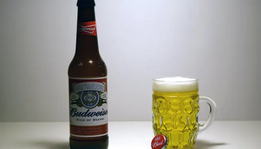 Butelka i kufel pełne złotego piwa Budweiser. Obok leży kapsel z napisem "Bud".