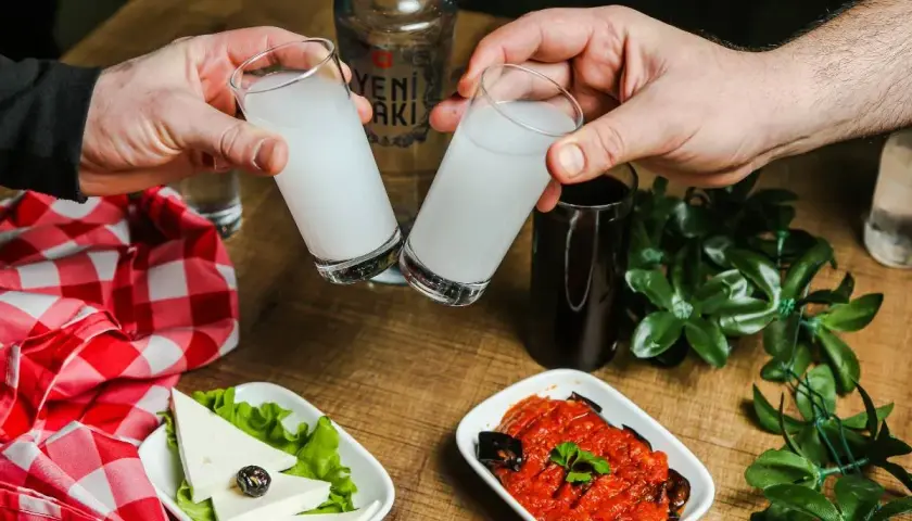 Dwie szklanki z mleczną wódką turecką (rakı) unoszą się do toastu. Na stole ser biały, oliwka i sos pomidorowy z bakłażanem.