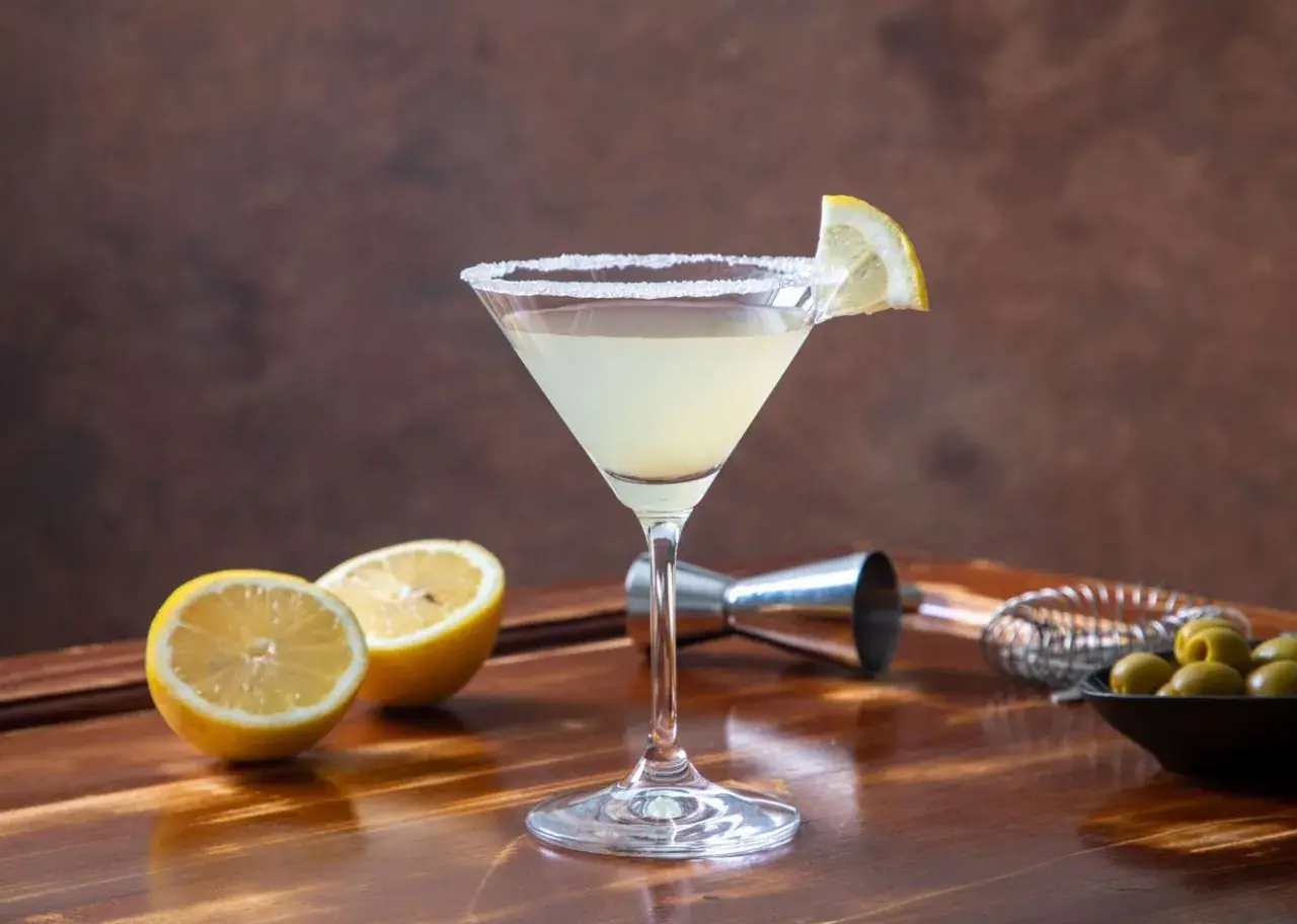 Klasyczny martini z cytryną i solą na kieliszku. W tle cytryny, oliwki i akcesoria barmańskie. Poznaj różne martini rodzaje.