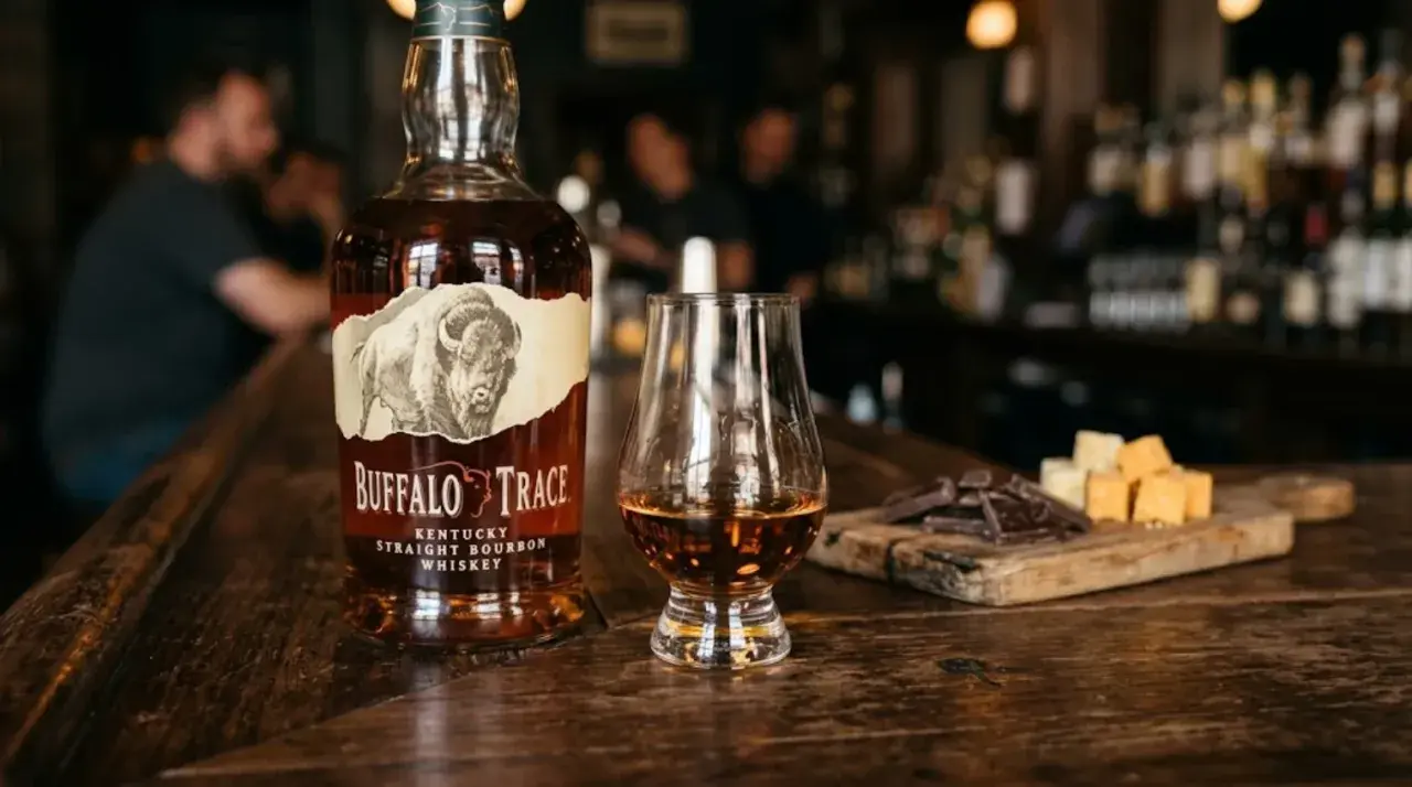 Butelka burbona Buffalo Trace, kieliszek z bursztynowym płynem, kawałki czekolady i sera na drewnianej desce.