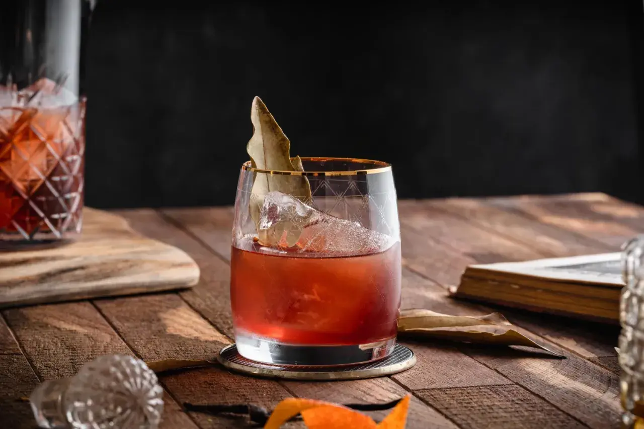 Elegancki koktajl, prawdopodobnie martini extra dry, z lodem i liściem laurowym, na drewnianym stole obok starej książki.
