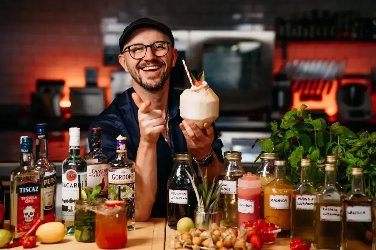 Uśmiechnięty barman z koktajlem w dłoni, otoczony butelkami alkoholi, w tym Bacardi. Sprawdź aktualną bacardi cena!
