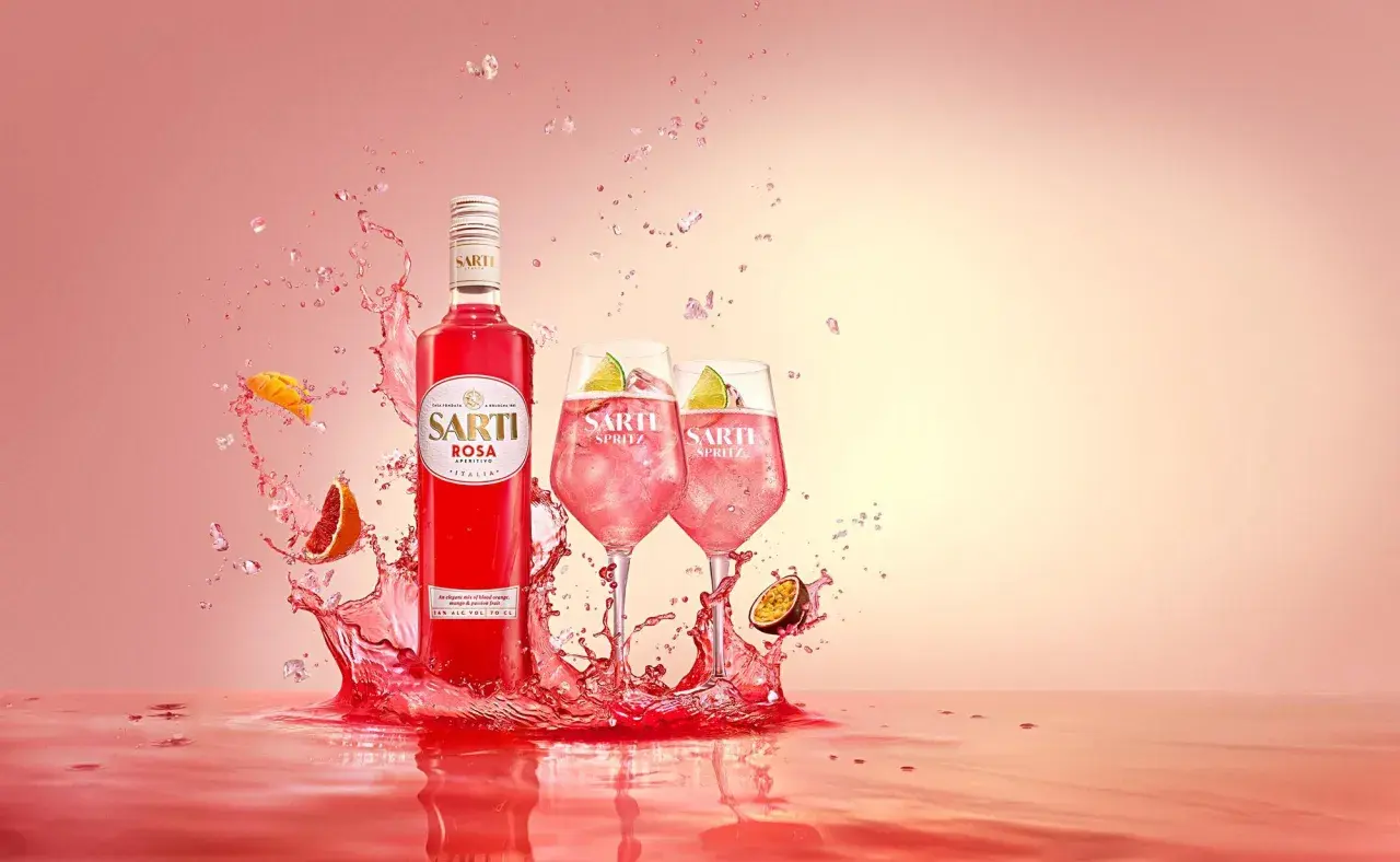 Sarti Rosa Aperitivo i dwa drinki Sarti Spritz z lodem i limonką, otoczone eksplozją owoców i różowego płynu.