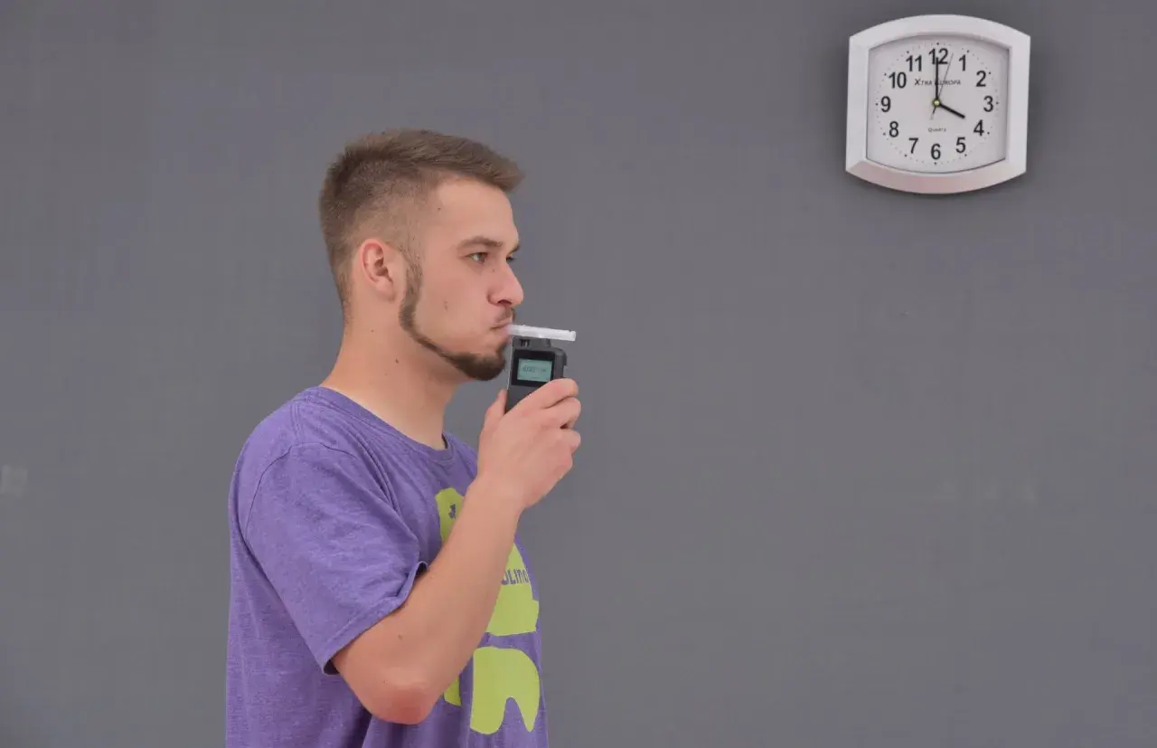 Mężczyzna dmucha w alkomat, obok wisi zegar.