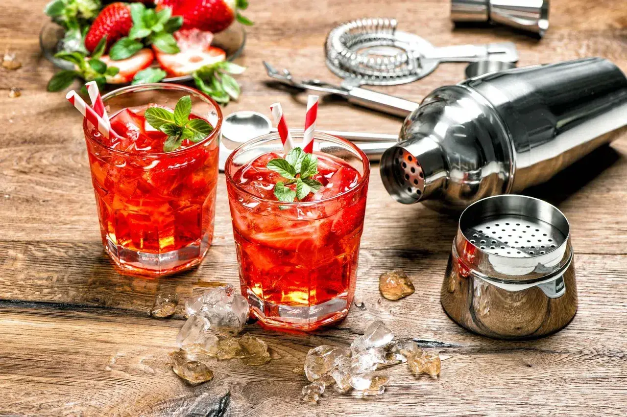 Dwa orzeźwiające drinki z martini rosato, lodem i truskawkami, obok shakera i akcesoriów barmańskich.