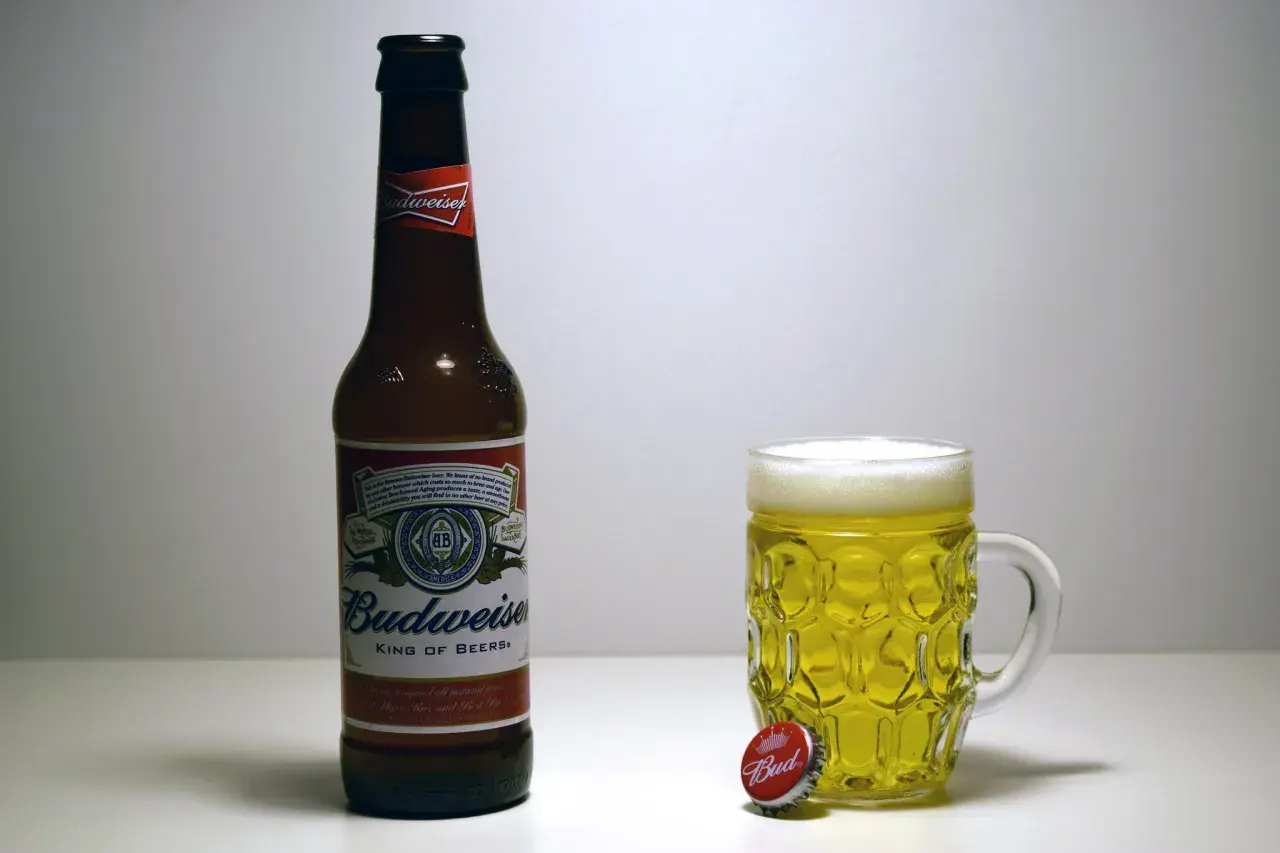 Butelka i kufel pełne złotego piwa Budweiser. Obok leży kapsel z napisem "Bud".