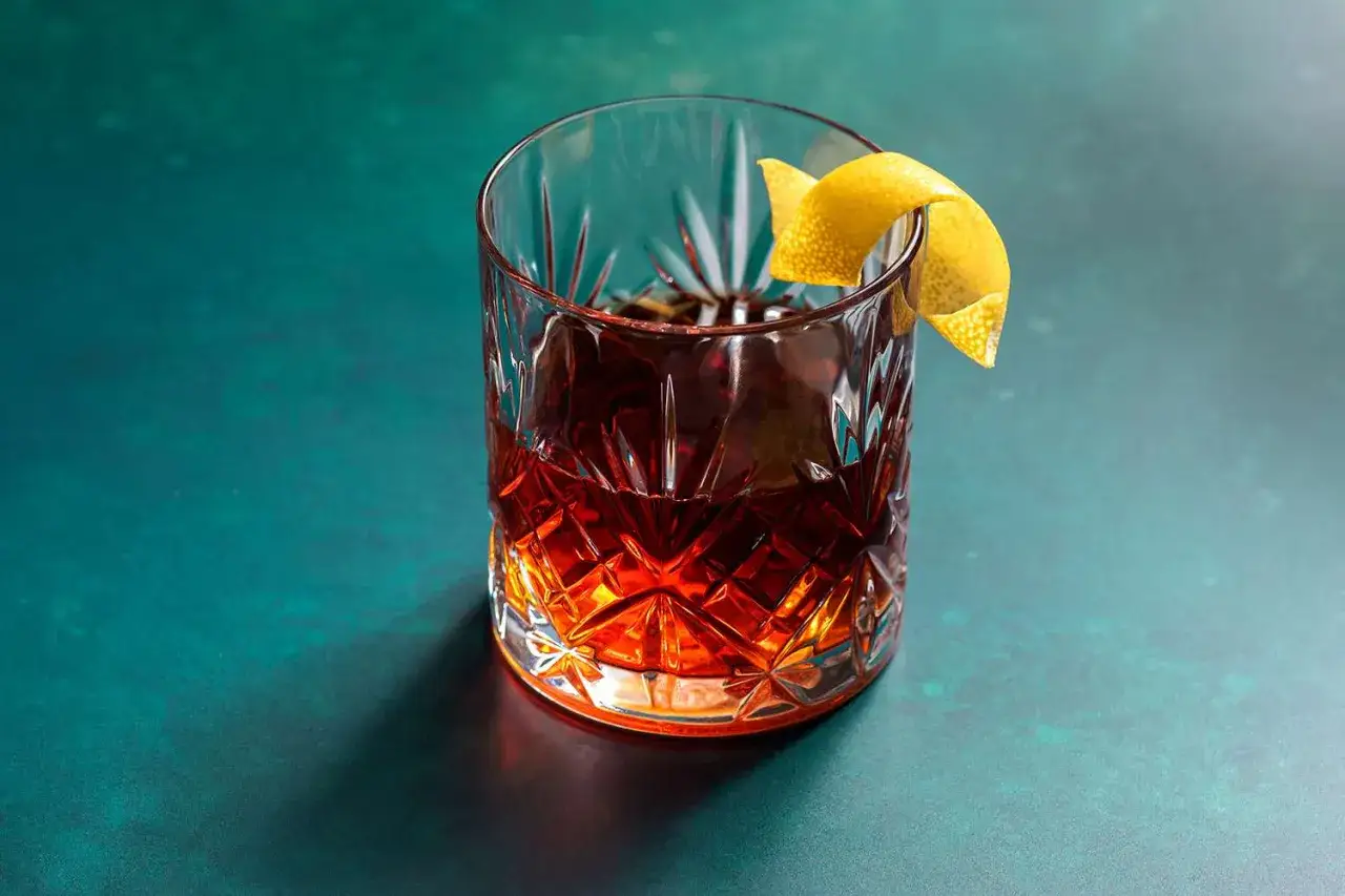 Klasyczny Sazerac w szklance typu old-fashioned, ozdobiony skórką cytryny, na ciemnozielonym tle.