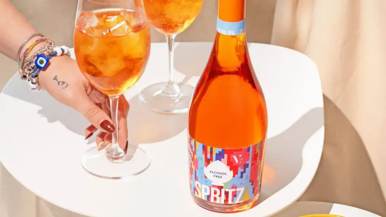 Ręka z bransoletkami sięga po kieliszek z drinkiem. Obok butelka aperol bezalkoholowy.