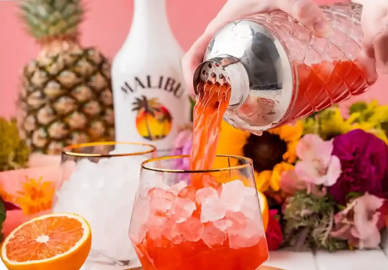 Malibu alkohol nalewany do szklanki z lodem, w tle ananas, pomarańcza i kwiaty.