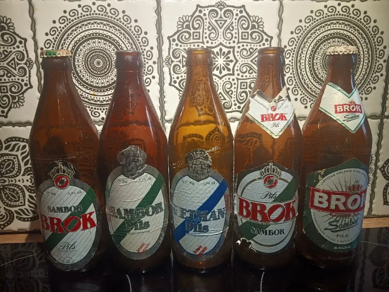 Pięć butelek piwa Brok Sambor Pils, z etykietami w kolorach zielonym, białym i czerwonym, na tle ozdobnych płytek.