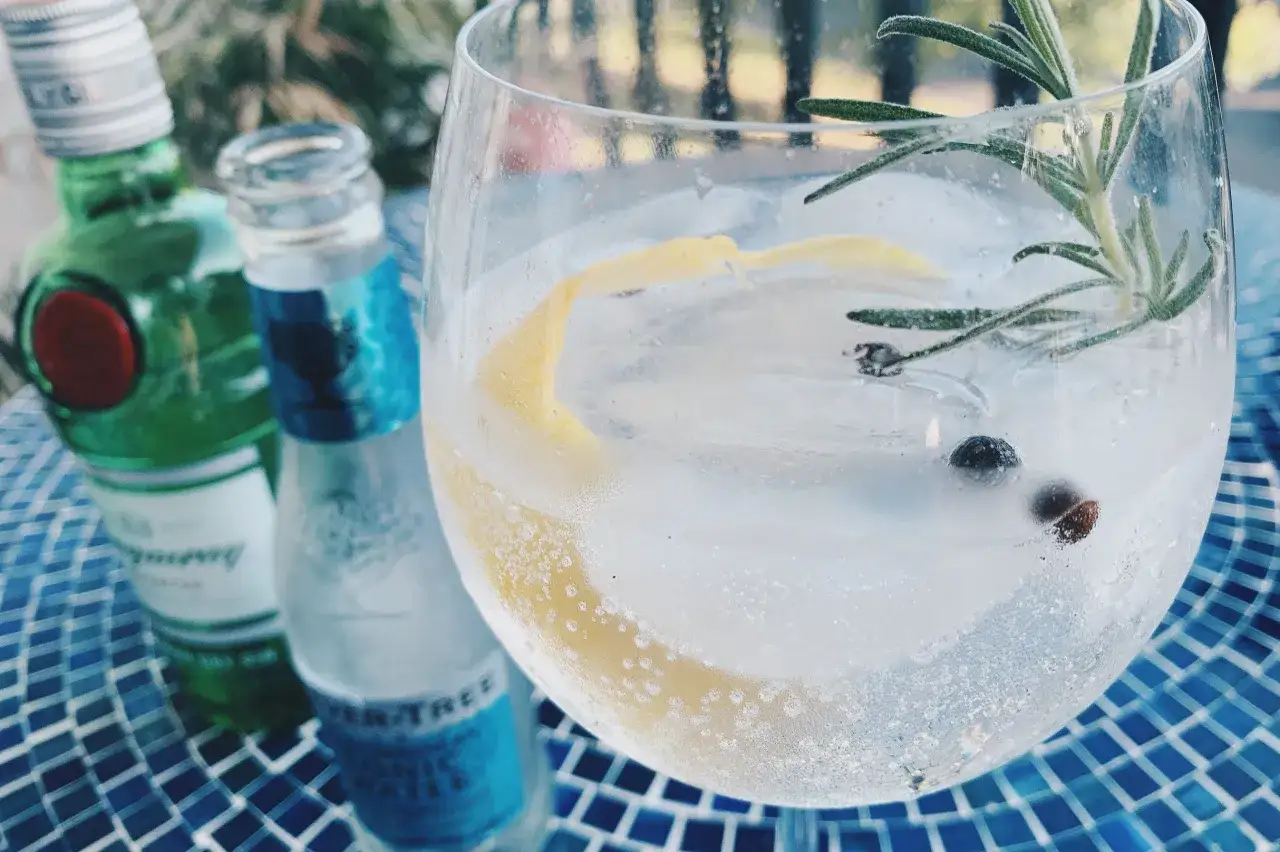Orzeźwiający gin z tonikiem, z cytrynową skórką, jagodami jałowca i rozmarynem, obok butelek Bombay Sapphire i Fever-Tree.