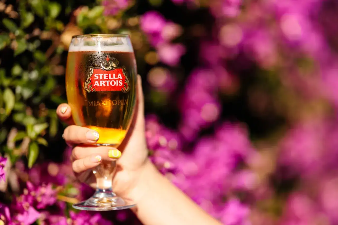 Dłoń trzyma kieliszek orzeźwiającego piwa Stella Artois z pianką, na tle kwitnących, fioletowych kwiatów.