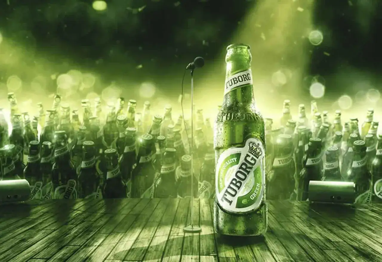 Zielona butelka piwa Tuborg Green na scenie, otoczona tłumem innych butelek. W tle mikrofon, sugerujący koncert.